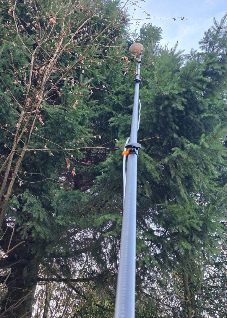 Ein langer, grauer Mast mit einer runden Spitze vor grünen Bäumen.